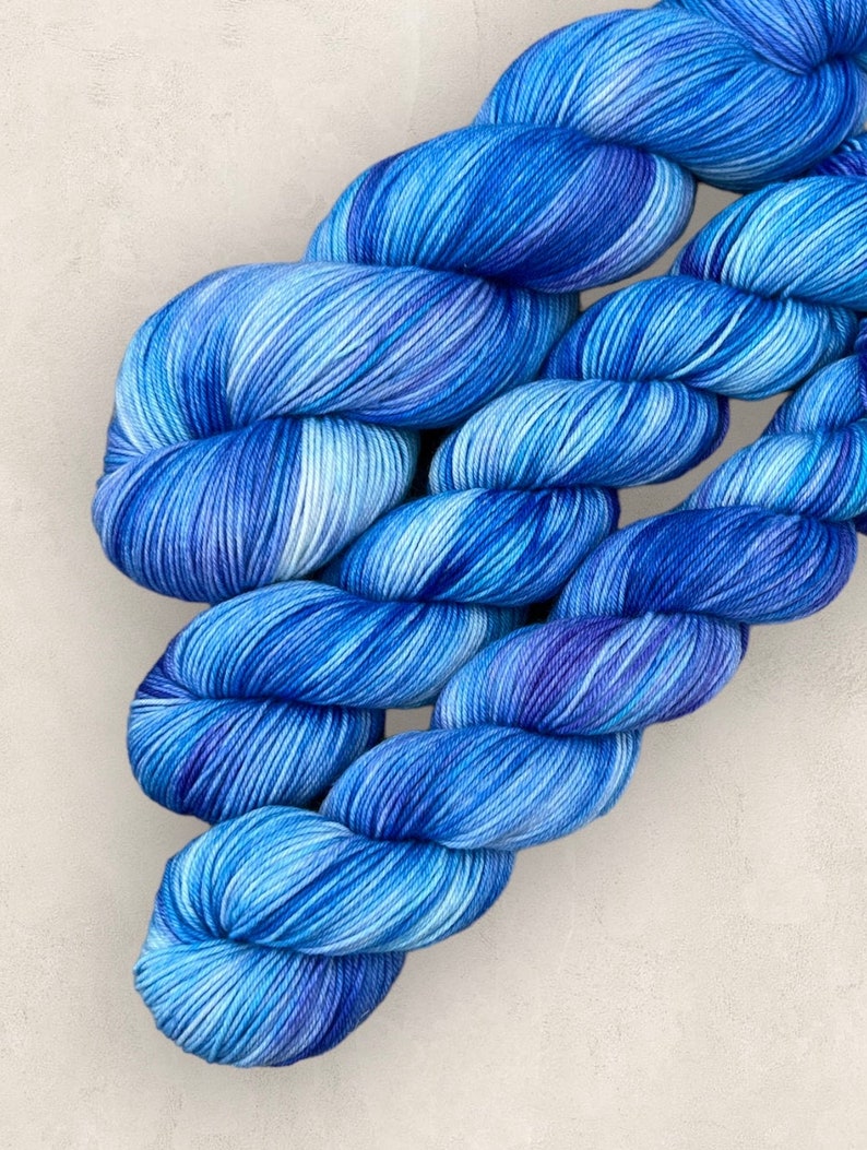 May include: A skein of blue hand-dyed yarn, with a gradient from light to dark indigo. The yarn is tightly twisted, displaying subtle colour and texture variations. Suitable for knitting or crochet.