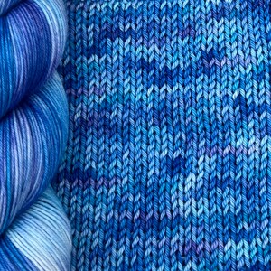 May include: A close-up of a skein of blue yarn next to a knitted fabric sample. The yarn is a gradient of blues and white. The knitted fabric is a textured pattern in shades of blue.