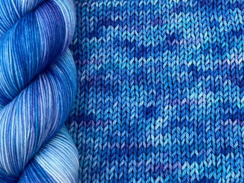 May include: A close-up of a skein of blue yarn next to a knitted fabric sample. The yarn is a gradient of blues and white. The knitted fabric is a textured pattern in shades of blue.