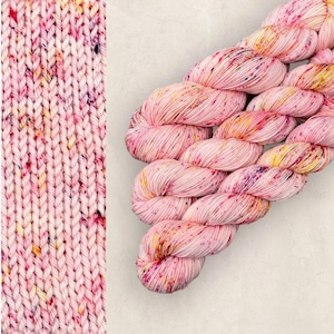 May include: Pink yarn skeins with colorful speckles. The yarn is a light pink color with flecks of yellow, red, and purple. The image also shows a close-up of the knitted fabric, showcasing the texture and color variations. Ideal for knitting and crochet projects.