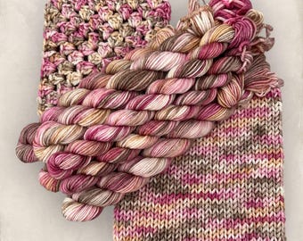 May include: An overhead shot showcases a collection of yarn and knitted items. The yarn is a mix of pink, brown, and cream colors. A crocheted square and a knitted piece are also visible, both featuring similar color palettes. The items suggest a theme of crafting and textile art.