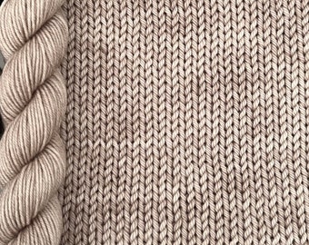 Könnte beinhalten: Nahaufnahme eines beigen gestrickten Stoffes mit einem Zopfmuster. Der Stoff liegt neben einer Garnrolle in Beige.