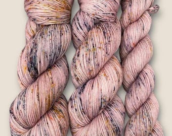 Op de afbeelding: Drie bollen roze garen met vlekken in paars, bruin en goud. Het garen is zacht en pluche, en het zou perfect zijn om een ​​gezellige trui of deken te breien of te haken.