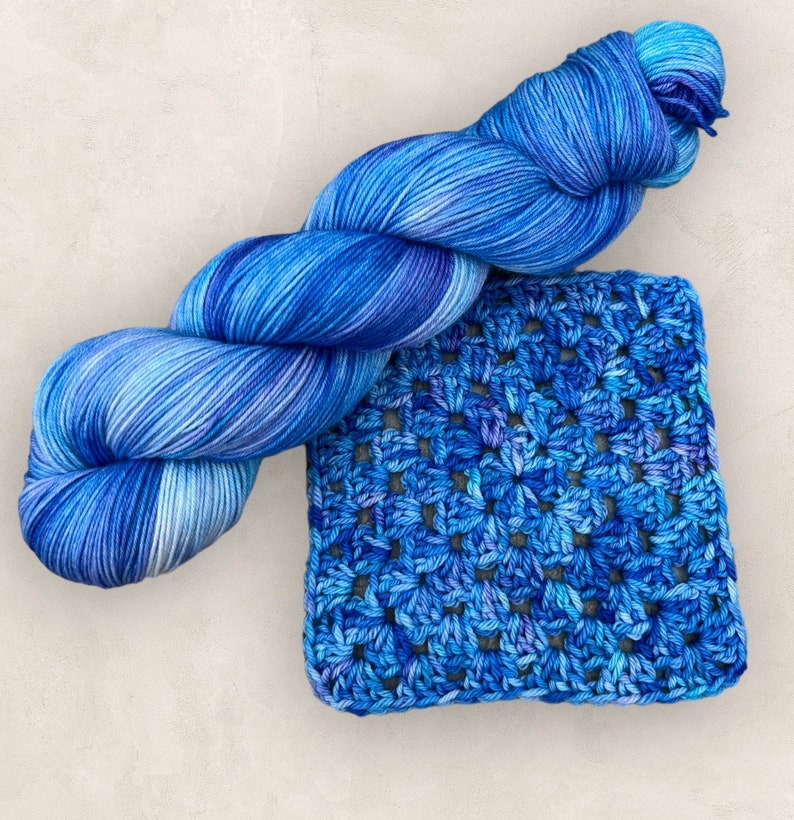 May include: A ball of blue variegated yarn and a crocheted square. The yarn is a mix of blues and white. The crocheted square features a floral pattern in various shades of blue.