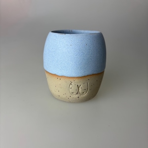 Könnte beinhalten: Handgefertigte Keramiktasse mit hellblauem Oberteil und beigefarbenem Unterteil. Der beige Bereich hat ein skurriles Design. Die Tasse hat eine abgerundete Form und eine gesprenkelte Textur, geeignet für Getränke.