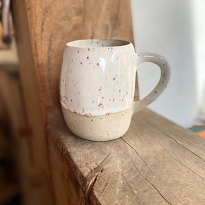 Könnte beinhalten: Eine weiße Keramiktasse mit braunen Flecken und einem gesprenkelten beigefarbenen Boden. Die Tasse hat einen abgerundeten Körper und einen Henkel.