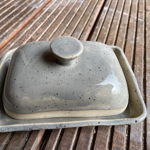 Könnte beinhalten: Eine graue Keramik-Butterdose mit Deckel. Die Dose hat eine gesprenkelte Glasur und einen weißen Rand.