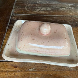 Könnte beinhalten: Eine rosa Keramik-Butterdose mit Deckel und einem gesprenkelten Überzug. Die Dose steht auf einer Holzoberfläche.
