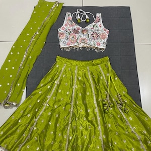May include: A lime green, three-piece Indian outfit. The set includes a sleeveless floral embroidered top, a matching skirt, and a sheer scarf. The skirt has a white floral border. The outfit is embellished with gold accents.