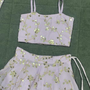 May include: A lavender-colored crop top and skirt set with gold floral embroidery and sequins. The top has thin straps and a square neckline. The skirt has a gathered waist and a flowing silhouette.