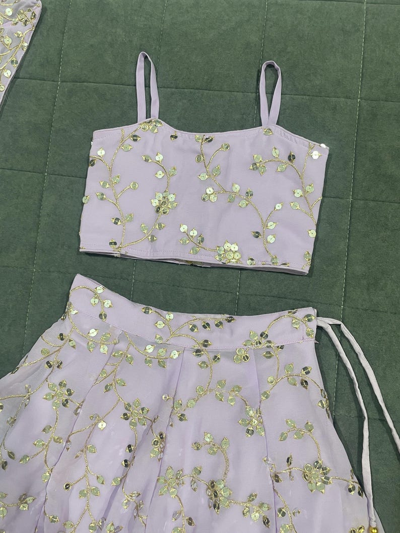 May include: A lavender-colored crop top and skirt set with gold floral embroidery and sequins. The top has thin straps and a square neckline. The skirt has a gathered waist and a flowing silhouette.