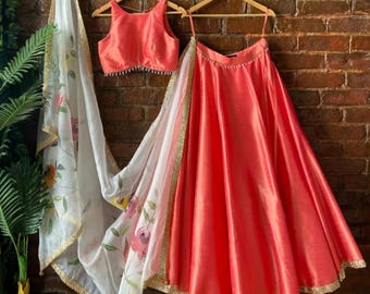 May include: A coral-colored sleeveless blouse and a matching full-length pleated skirt, paired with a sheer white dupatta featuring a floral print and gold-toned trim. The set is displayed on two wooden hangers against a rustic brick wall.