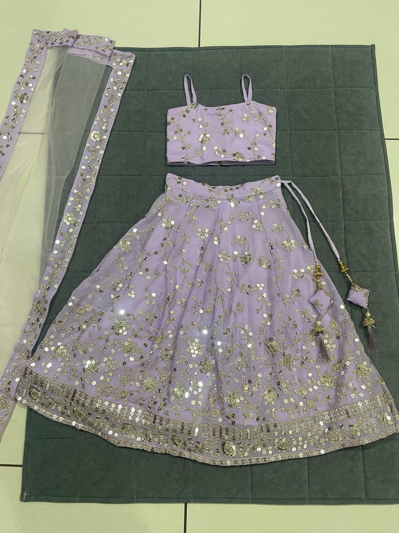 May include: A lavender girls' outfit with a sleeveless top and a skirt. The top and skirt are decorated with silver sequins and embroidery. The outfit also includes a sheer lavender dupatta.