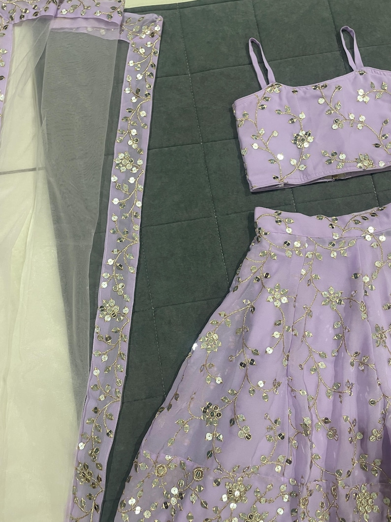 May include: A lilac-colored three-piece outfit with silver embroidery. The outfit includes a sleeveless top, a skirt, and a sheer dupatta with a silver border.