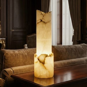 Puede incluir: Lámpara rectangular con aspecto natural y texturizado. La lámpara emite una luz cálida y brillante, y está hecha de un material claro con vetas marrones y beige. Se encuentra sobre una mesa de madera oscura, un elemento decorativo.