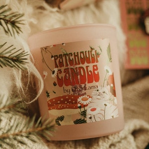 Puede incluir: Una vela rosa con una etiqueta blanca que dice "Patchouli Candle by Dzo Lama". La etiqueta presenta un gato blanco, hongos y flores.