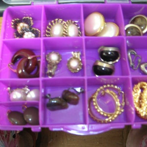 May include: A purple plastic storage box with compartments filled with various earrings, including pearl, gold, and black earrings.