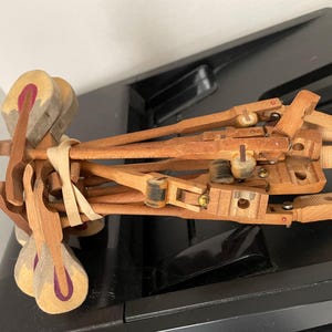 May include: A collection of wooden piano action parts, including hammers with felt heads and purple centers, connected by a rubber band. The mechanism features light brown wooden shanks, knuckles, and flanges, showing intricate mechanical components.