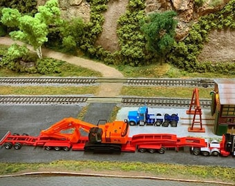 May include: A model train set with a red and orange truck hauling a large excavator. The truck is connected to a flatbed trailer with a crane arm. The truck is parked next to a small building with a brown roof and a blue door. The building has a sign that says "W.S.S."