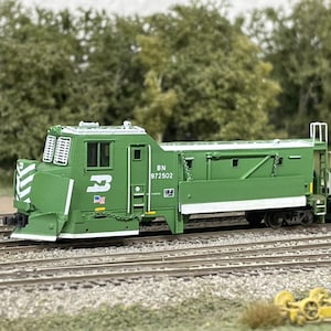 May include: A model train locomotive in green, displaying the number "3" and the letters "BN" and the number "972502". The model train is on a miniature railway track, with a red and yellow train carriage visible in the background.