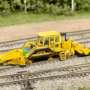 Puede incluir: Una máquina de mantenimiento de vías de ferrocarril modelo en amarillo con un detalle negro y verde en el lateral. La máquina está en una vía de ferrocarril modelo.