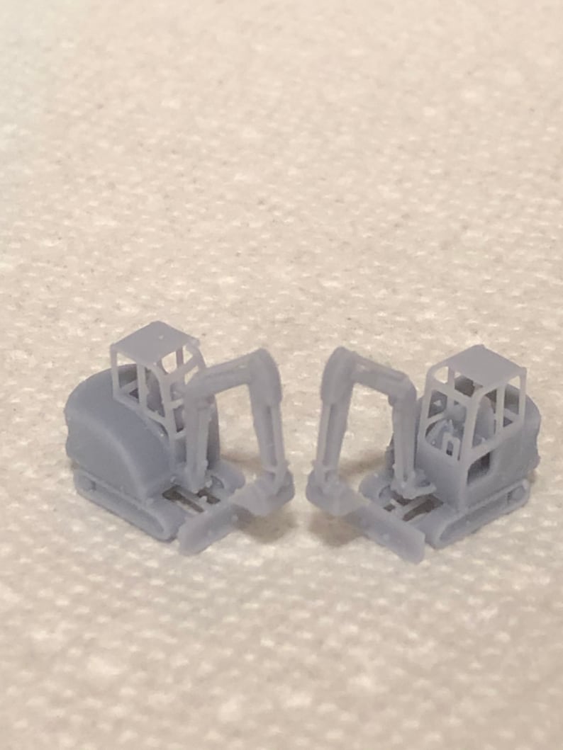 May include: Two small, grey, plastic toy excavators with black tracks and a detailed design. The excavators are facing each other with their arms extended.
