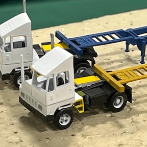 May include: Miniature white semi-trucks with white cabs and black chassis. One truck has a blue trailer, and the other has a yellow trailer. The trucks are on a tan surface.