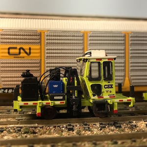 Puede incluir: Un vagón de ferrocarril modelo amarillo y negro con un logotipo de CSX y una pequeña cabina. El vagón tiene un parachoques a rayas blanco y negro y un techo a rayas blanco y negro. El vagón está en una vía de ferrocarril modelo.