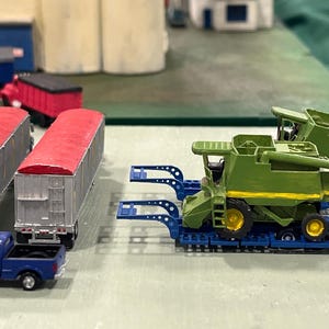 Puede incluir: Vehículos agrícolas en miniatura, incluyendo una camioneta azul tirando de dos remolques plateados con techos rojos, y dos cosechadoras verdes sobre remolques azules. Otros edificios y vehículos se ven al fondo.