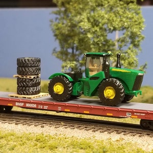 May include: A green tractor with black tires is being transported on a flatbed railroad car. The tractor is secured to the car with straps. A stack of tires is also on the car. The car is labeled "BNSF 800036".