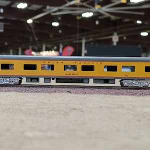 Pode incluir: Um vagão de trem modelo amarelo e cinza com as palavras "UNION PACIFIC" e "LAKE FOREST" impressas no lado. O vagão tem várias janelas e está sobre um trilho.