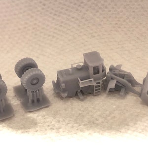 May include: A grey plastic model of a construction vehicle with a bucket, a cab, and four wheels. The model is disassembled into five pieces, including the cab, the bucket, and two sets of wheels.