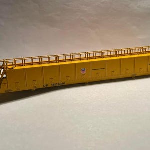 May include: A yellow model train car with a detailed design. The car has a rectangular shape with a long, flat top and a series of vertical panels. The words "UP 980354MW" are printed on the side, along with the Union Pacific logo.