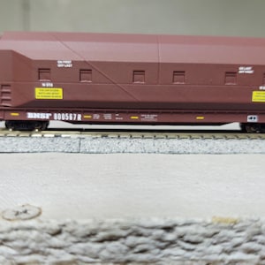 Op de afbeelding: Een bruine modeltreinwagen met de tekst "BNSF 000567R" erop gedrukt. De wagen heeft een geel waarschuwingslabel met zwarte tekst. De wagen staat op een grijs spoor.