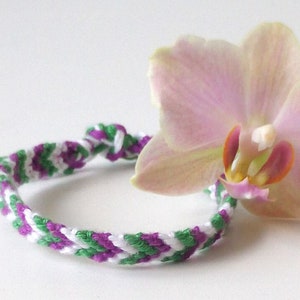 Könnte beinhalten: Ein weißes, grünes und violettes Freundschaftsarmband mit einer rosa Orchideenblüte darauf.
