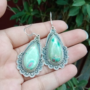Peut inclure: Une paire de boucles d'oreilles en argent avec des gouttes de pierres précieuses vertes et roses. Les boucles d'oreilles ont un design délicat et complexe.