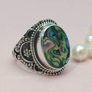 Pode incluir: Um anel de prata com uma incrustação oval de concha de abalone. O anel tem um design detalhado e ornamentado com uma borda de corda e pequenos acentos redondos.