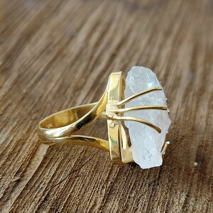 May include: A gold ring with a clear quartz crystal set in a gold cage. The ring is on a wooden surface.