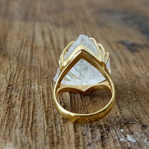 May include: A gold ring with a clear quartz crystal set in a cage-like design. The ring is shown from the back, revealing the gold band.