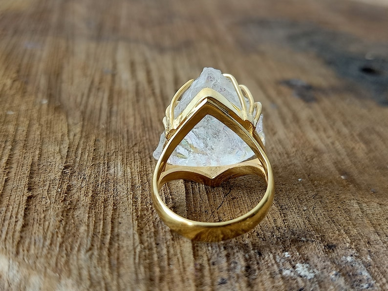 May include: A gold ring with a clear quartz crystal set in a cage-like design. The ring is shown from the back, revealing the gold band.