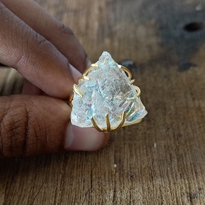 May include: A clear, iridescent crystal ring with a gold wire setting. The crystal is shaped like a triangle and has a rough, natural texture.