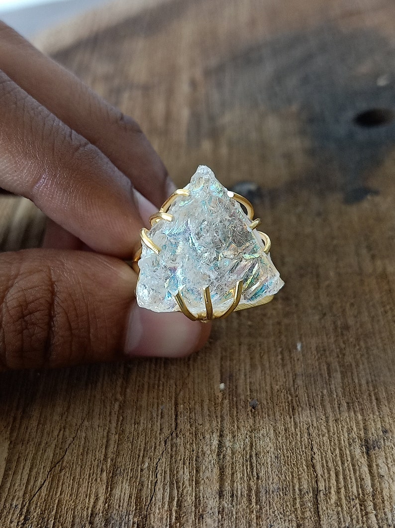 May include: A clear, iridescent crystal ring with a gold wire setting. The crystal is shaped like a triangle and has a rough, natural texture.