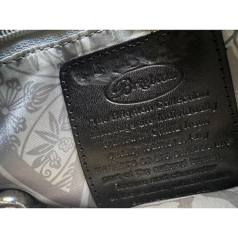 Puede incluir: Primer plano de una etiqueta de cuero negro con el logotipo de Brighton y texto. La etiqueta est&aacute; cosida a una bolsa con un forro estampado floral. El texto describe la calidad y la belleza natural del cuero.