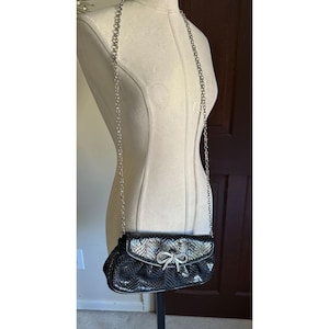 Puede incluir: Bolso de noche negro y plateado con correa de cadena. El bolso presenta un patr&oacute;n texturizado, similar a una serpiente, y un cierre de lazo plateado. El bolso se exhibe en un maniqu&iacute;.
