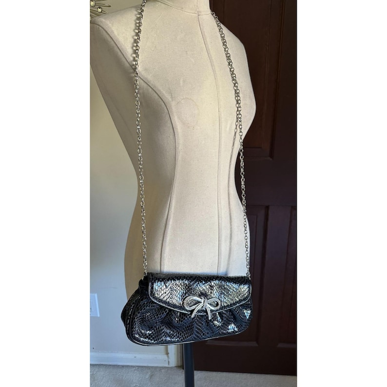 Puede incluir: Bolso de noche negro y plateado con correa de cadena. El bolso presenta un patr&oacute;n texturizado, similar a una serpiente, y un cierre de lazo plateado. El bolso se exhibe en un maniqu&iacute;.