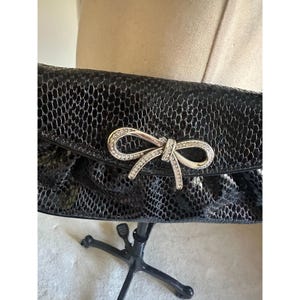 Puede incluir: Bolso de mano negro con un patr&oacute;n texturizado similar a la piel de serpiente. La solapa presenta un lazo plateado adornado con peque&ntilde;os y brillantes pedrer&iacute;a. Un accesorio elegante para ocasiones especiales.