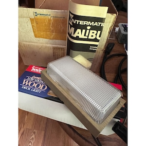 May include: A white, rectangular, low voltage deck light with a wood frame. The light is packaged in a box with the text "Intermatic Malibu Compact Low Voltage Wood Deck Light" printed on it.