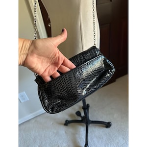 Puede incluir: Bolso de mano negro con correa de cadena. El bolso tiene un estampado de piel de serpiente y un cierre de solapa. La cadena es de color plateado. El bolso se exhibe en un soporte de metal negro.
