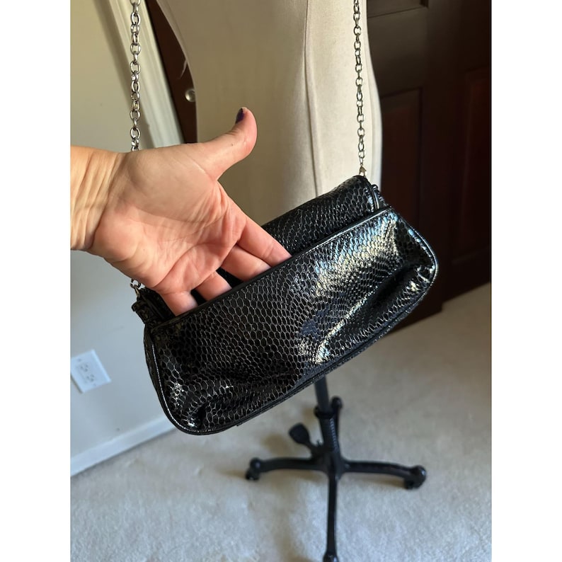 Puede incluir: Bolso de mano negro con correa de cadena. El bolso tiene un estampado de piel de serpiente y un cierre de solapa. La cadena es de color plateado. El bolso se exhibe en un soporte de metal negro.