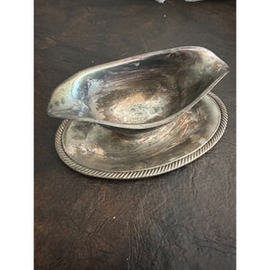 May include: A silver gravy boat with a matching saucer. The boat has a rounded shape and a lip on one side. The saucer has a rope-like design around the edge.